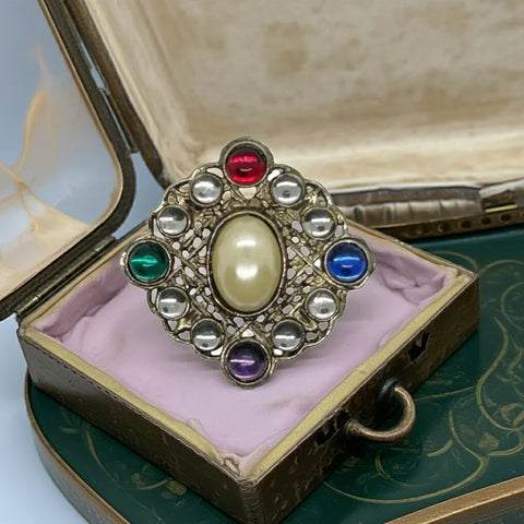 This striking brooch boasts a majestic diamond design, crafted from intricately detailed filigree metal and adorned with a sizable oval faux pearl at its heart. Surrounding the pin are lustrous round cabochons in a rich palette of red, green, purple, and blue, elegantly interspersed with two clear gems between each color
