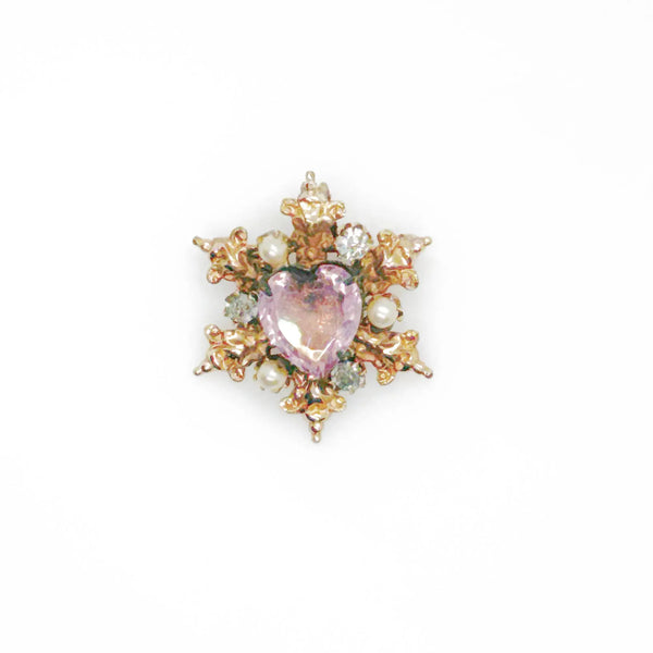 A gold-toned brooch with a central heart-shaped, light purple gemstone. The brooch is surrounded by ornate gold filigree, small pearls, and clear crystals, creating a vintage, elegant design.