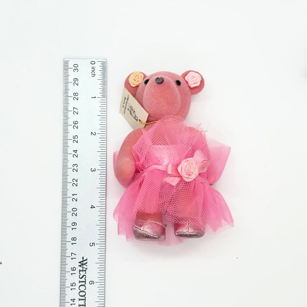 A small pink teddy bear figurine wearing a pink tulle dress with a fabric rose, shown next to a metal ruler for scale on a white background.