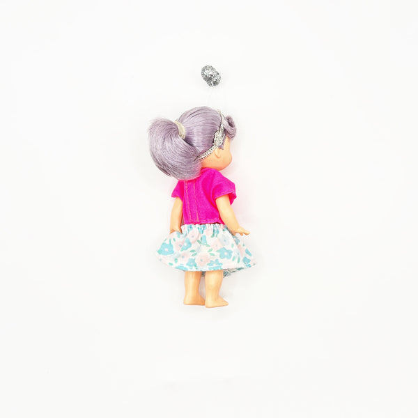 Rear view of a 4-inch Mattel Kelly Club doll with grey ponytail hair and a silver headband. The collectible doll is dressed in a vibrant pink short-sleeve shirt and a blue floral print skirt on a clean white backdrop