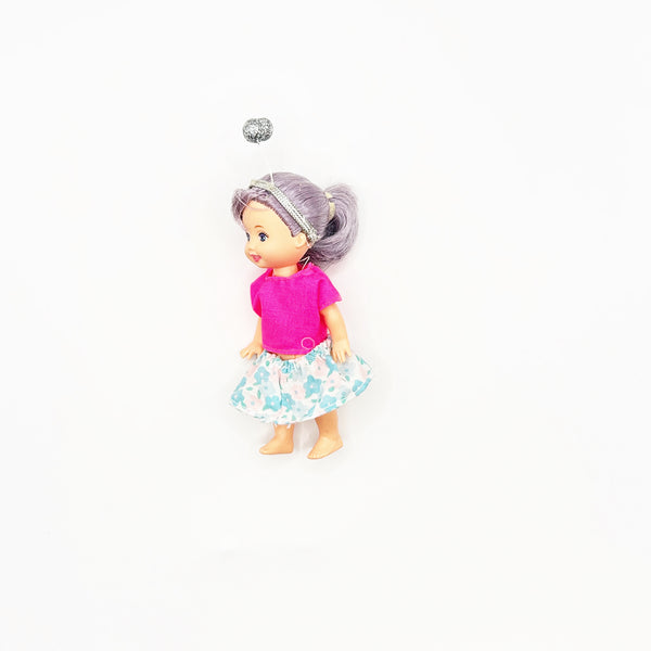 A vintage 4-inch Mattel Barbie Kelly Club mini doll with silvery-purple hair styled in a high side ponytail and a silver glitter headband. The doll is wearing a vibrant pink short-sleeve top and a white flared skirt with a teal and light-blue abstract floral print, positioned against a seamless white