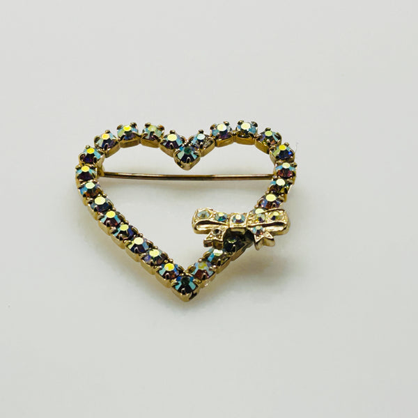 This charming mid-century brooch captures all the sparkle and romance of vintage glamour. The delicate gold-tone heart showcases shimmering aurora borealis rhinestones that dance with rainbow fire, while a sweet bow accent adds an extra touch of whimsy.