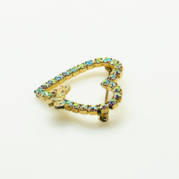 This charming mid-century brooch captures all the sparkle and romance of vintage glamour. The delicate gold-tone heart showcases shimmering aurora borealis rhinestones that dance with rainbow fire, while a sweet bow accent adds an extra touch of whimsy.