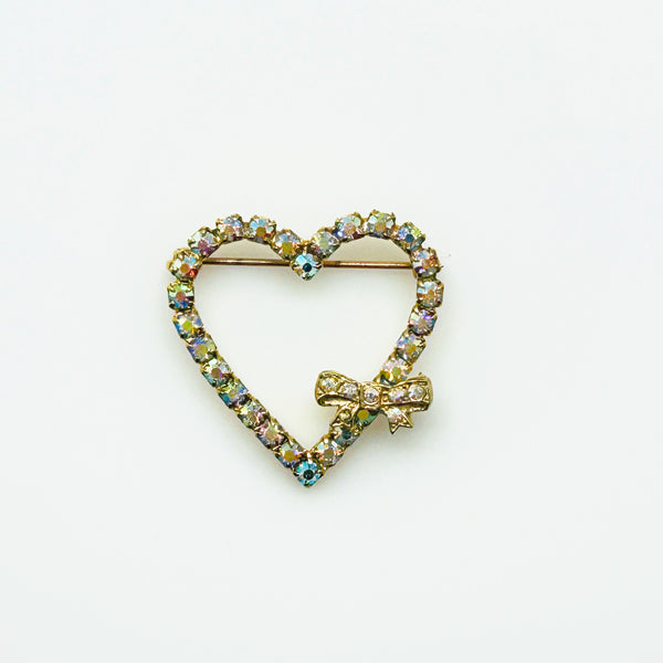 This charming mid-century brooch captures all the sparkle and romance of vintage glamour. The delicate gold-tone heart showcases shimmering aurora borealis rhinestones that dance with rainbow fire, while a sweet bow accent adds an extra touch of whimsy.