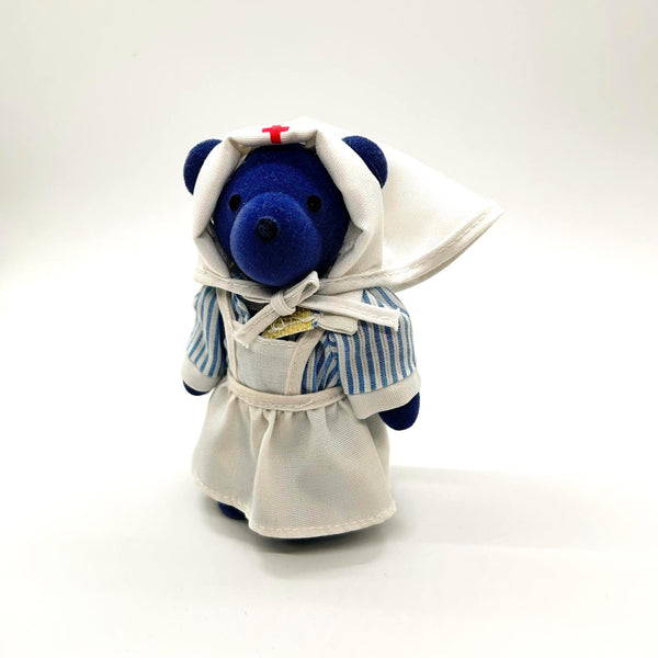 A small plush teddy bear dressed as a nurse, with a blue body, white apron and head covering, and a small red cross on the hat, photographed on a clean white background.
