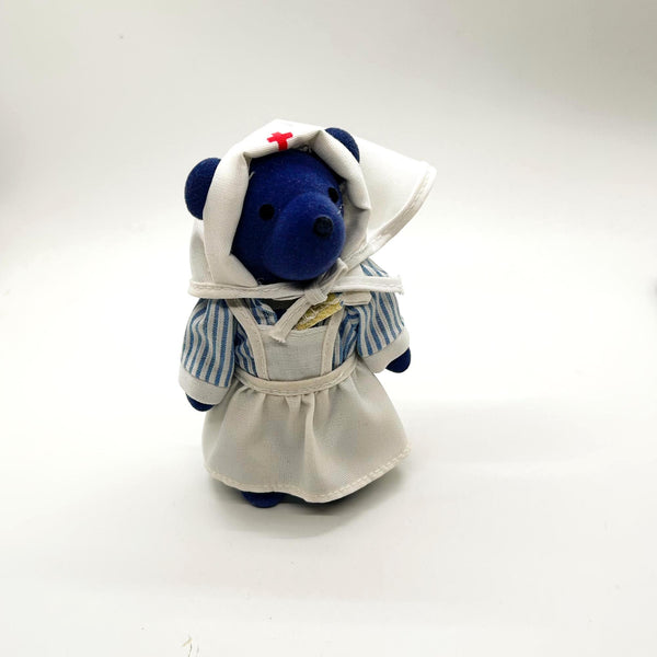 A small plush teddy bear dressed as a nurse, featuring a blue body, a white apron and head covering with a red cross, and blue-and-white striped sleeves, photographed on a plain white background.