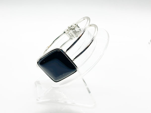 This striking vintage bracelet showcases a bold black square onyx stone set in polished silver. The architectural design features dual silver bands converging at the center stone with a hinged hook-bangle closure for easy wear. 