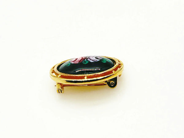 Picture a charming piece of wearable nostalgia: this oval brooch pairs a moody black canvas with cheerful painted florals, all framed in warm gold-tone metal. It's a mid-20th century take on Victorian mourning jewelry aesthetics
