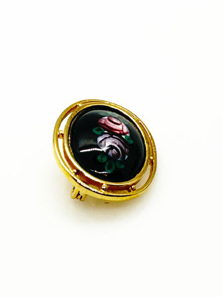 Picture a charming piece of wearable nostalgia: this oval brooch pairs a moody black canvas with cheerful painted florals, all framed in warm gold-tone metal. It's a mid-20th century take on Victorian mourning jewelry aesthetics