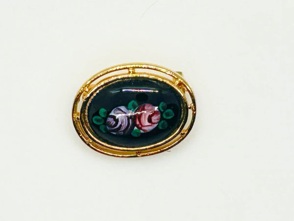 Picture a charming piece of wearable nostalgia: this oval brooch pairs a moody black canvas with cheerful painted florals, all framed in warm gold-tone metal. It's a mid-20th century take on Victorian mourning jewelry aesthetics