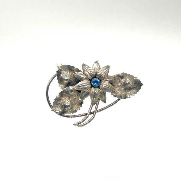 A vintage-style silver-toned flower brooch with textured leaves and a central round blue gemstone, displayed on a clean white background.