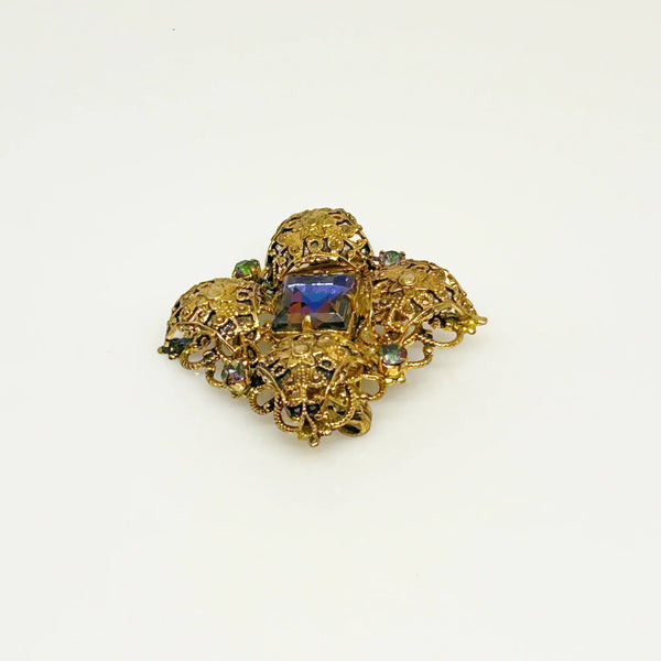 This exquisite brooch with an Old World aesthetic showcases finely crafted gold filigree petals encircling a square stone in elegant blue-purple tones. Between each delicate petal rests a round, iridescent stone that complements the central hues, adding subtle depth and shimmer. 