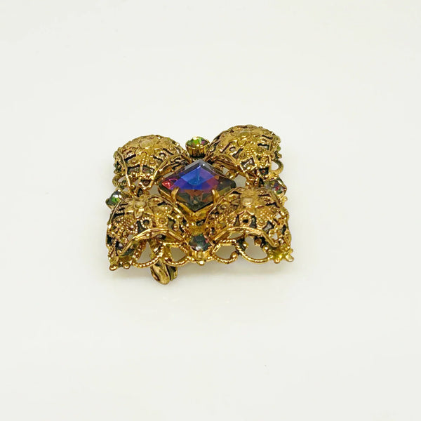 This exquisite brooch with an Old World aesthetic showcases finely crafted gold filigree petals encircling a square stone in elegant blue-purple tones. Between each delicate petal rests a round, iridescent stone that complements the central hues, adding subtle depth and shimmer. 