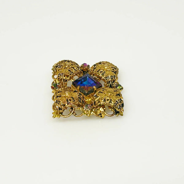 This exquisite brooch with an Old World aesthetic showcases finely crafted gold filigree petals encircling a square stone in elegant blue-purple tones. Between each delicate petal rests a round, iridescent stone that complements the central hues, adding subtle depth and shimmer. 