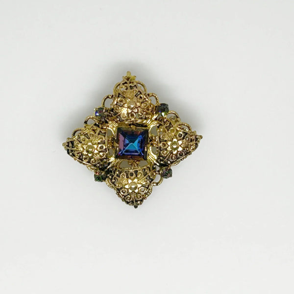 This exquisite brooch with an Old World aesthetic showcases finely crafted gold filigree petals encircling a square stone in elegant blue-purple tones. Between each delicate petal rests a round, iridescent stone that complements the central hues, adding subtle depth and shimmer. 