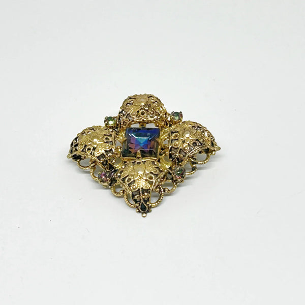 This exquisite brooch with an Old World aesthetic showcases finely crafted gold filigree petals encircling a square stone in elegant blue-purple tones. Between each delicate petal rests a round, iridescent stone that complements the central hues, adding subtle depth and shimmer. 