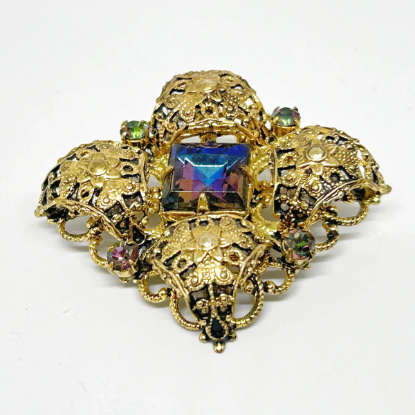 This exquisite brooch with an Old World aesthetic showcases finely crafted gold filigree petals encircling a square stone in elegant blue-purple tones. Between each delicate petal rests a round, iridescent stone that complements the central hues, adding subtle depth and shimmer. 