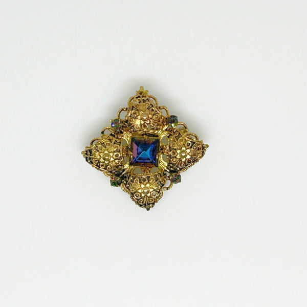 This exquisite brooch with an Old World aesthetic showcases finely crafted gold filigree petals encircling a square stone in elegant blue-purple tones. Between each delicate petal rests a round, iridescent stone that complements the central hues, adding subtle depth and shimmer. 