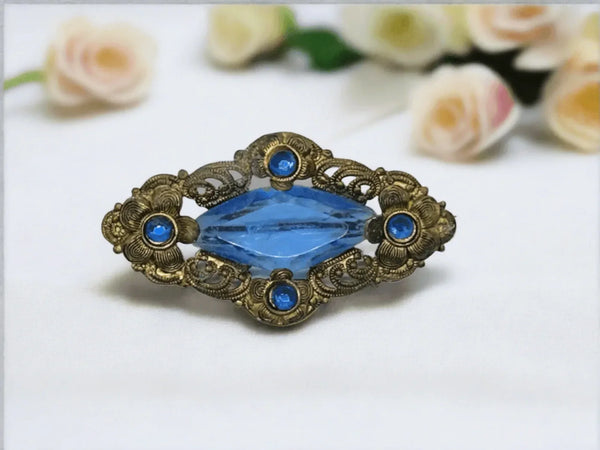 Step back in time with this striking antique Victorian brooch, showcasing intricate designs that will surely catch your eye. It boasts a brilliant clear blue stone in a bold square shape, surrounded by a delicate arrangement of four flower-shaped clusters with a dazzling blue round stone at its core, adorned with elegant swirls and scallops. 