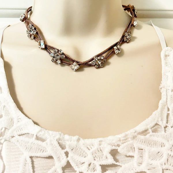 This vintage rhinestone necklace stands out immediately with diamond-princess cut rhinestones and clusters of round white rhinestones in alternating stations, all of which are prong-set and luminous. Two strands of rose gold tone metal are braided together and feature a hook clasp, measuring 15" in length. Minor signs of wear may be visible upon close inspection.