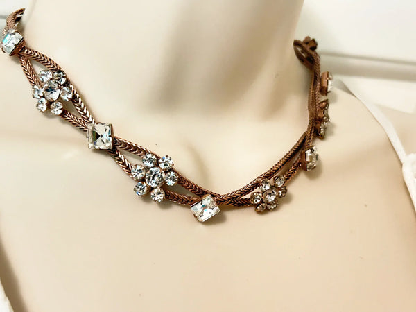 This vintage rhinestone necklace stands out immediately with diamond-princess cut rhinestones and clusters of round white rhinestones in alternating stations, all of which are prong-set and luminous. Two strands of rose gold tone metal are braided together and feature a hook clasp, measuring 15" in length. Minor signs of wear may be visible upon close inspection.