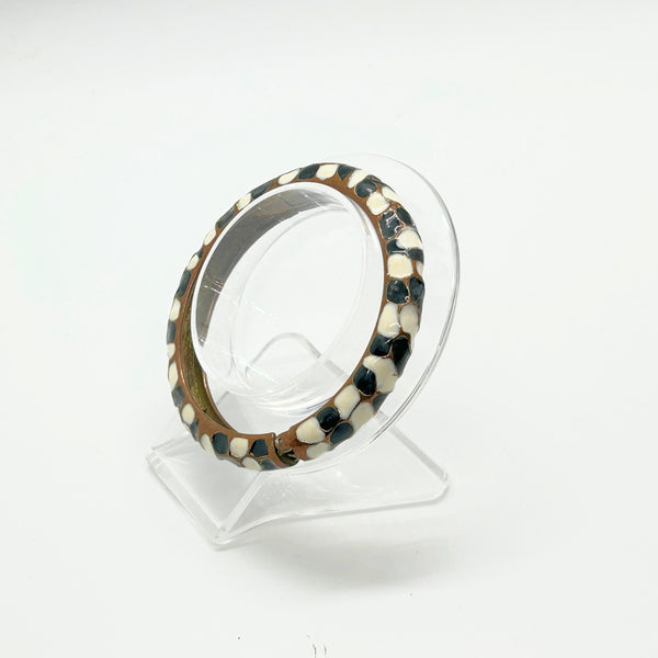 This striking hinge bangle bracelet showcases bold geometric patterns rendered in contrasting black and white enamel, accented with warm copper metalwork. The repeating abstract design creates visual interest from every angle, making it a versatile piece that complements both casual and dressed-up looks