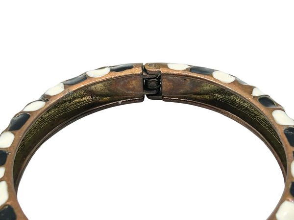 This striking hinge bangle bracelet showcases bold geometric patterns rendered in contrasting black and white enamel, accented with warm copper metalwork. The repeating abstract design creates visual interest from every angle, making it a versatile piece that complements both casual and dressed-up looks