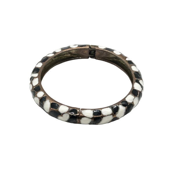 This striking hinge bangle bracelet showcases bold geometric patterns rendered in contrasting black and white enamel, accented with warm copper metalwork. The repeating abstract design creates visual interest from every angle, making it a versatile piece that complements both casual and dressed-up looks