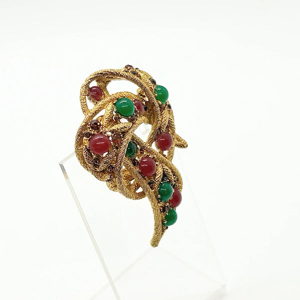 A vintage-style gold filigree brooch featuring red and green cabochon gemstones, displayed on a clear acrylic stand against a white background.