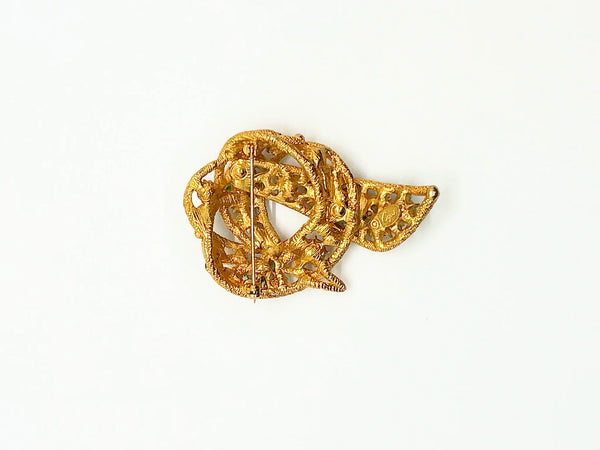 An image showing the reverse side of a textured gold-tone vintage costume jewelry brooch. The brooch is shaped like a textured, openwork abstract ribbon loop or bow with a dangling end pointing downward. A long, horizontal metal pin stem and safety clasp run across the upper back portion. On the lower section of the brooch, an oval cartouche containing the Capri maker's mark is stamped directly onto the textured filigree-style metal framework