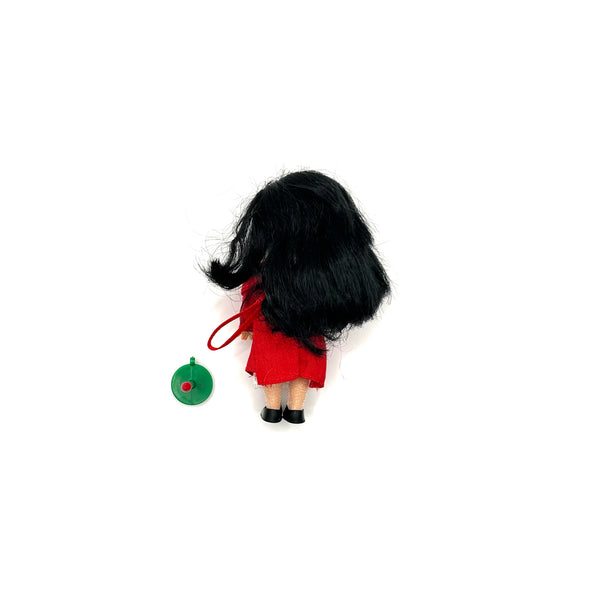 Rear view of a vintage Mattel Barbie Kelly Club Caroling doll ornament featuring thick black hair, a red dress, and black shoes, standing next to a green wreath accessory against a white background