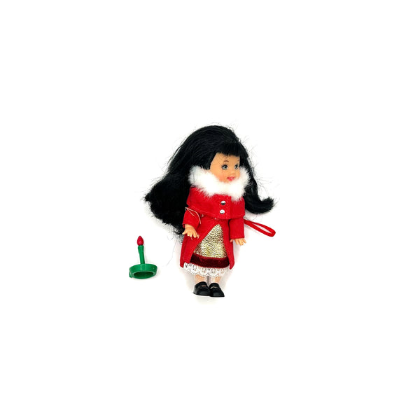 A loose 2001 Barbie Kelly Club Caroling Lorena holiday mini doll. The figure features long straight black hair, a bright red coat with white faux fur trim, a gold metallic patterned skirt, and small black shoes. Standing next to her is a miniature green plastic candle holder accessory
