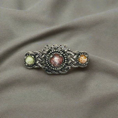 Embellish your style with the elegant and enduring Vintage Celtic Knot Style Brooch Art Glass. This beautifully crafted brooch features textured metal encasing stunning round stones in shades of pale green, red, and gold-amber.