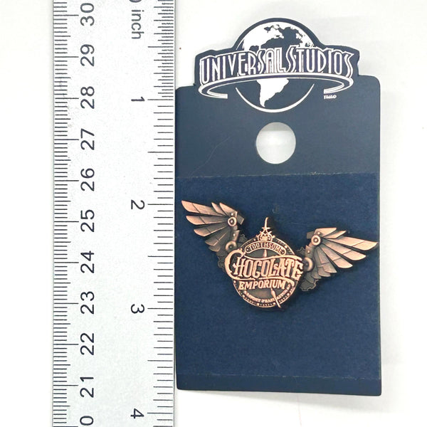 Official Universal Studios Parks Toothsome Chocolate Emporium steampunk logo lapel pin with mechanical wings, mounted on its original dark blue backing card.