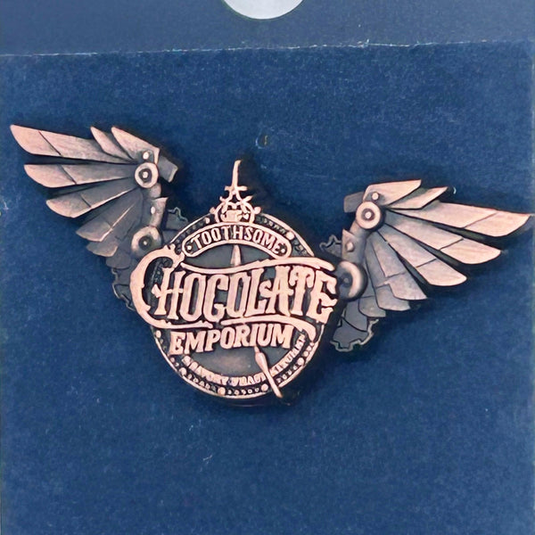 Official Universal Studios Parks Toothsome Chocolate Emporium steampunk logo lapel pin with mechanical wings, mounted on its original dark blue backing card.