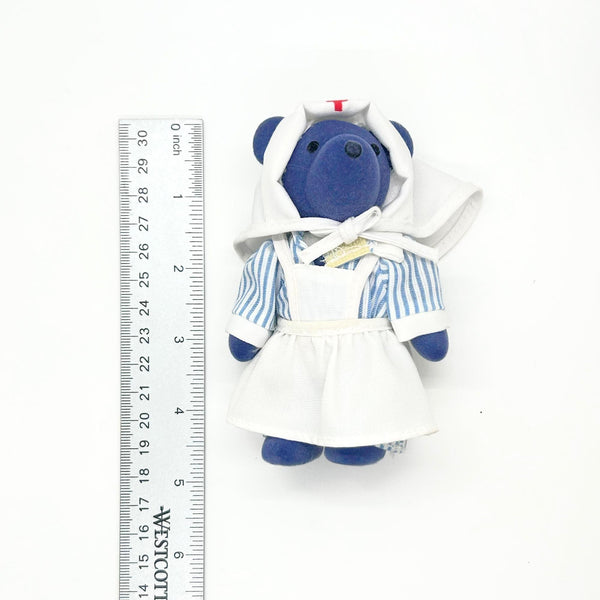 A small plush teddy bear dressed in a blue-and-white striped shirt, white apron, and nurse hat, shown next to a measuring ruler on a plain white background.