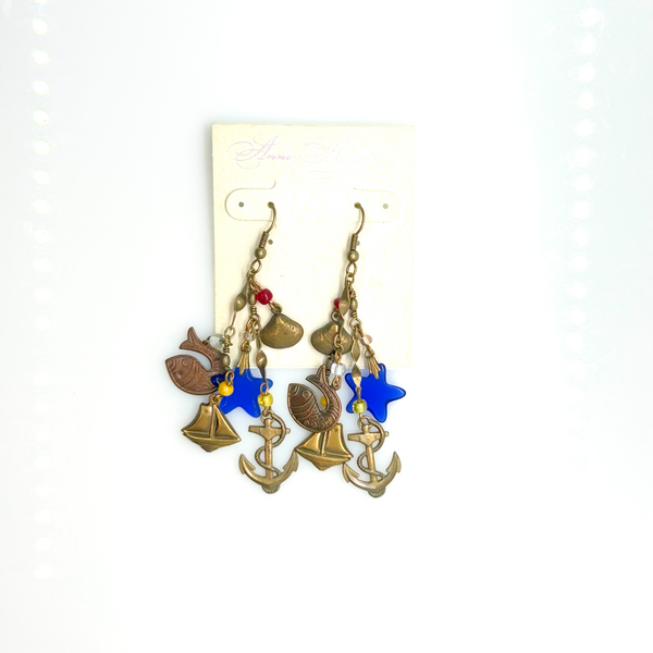 Vintage Cookie Lee nautical charm dangle earrings in antiqued brass with anchor, sailboat, fish, and blue star charms on a cream card.