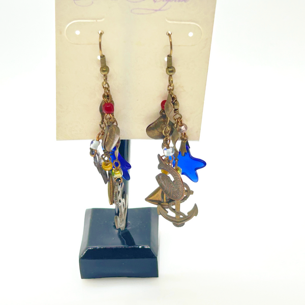 Vintage Cookie Lee nautical charm dangle earrings in antiqued brass with anchor, sailboat, fish, and blue star charms on a cream card.