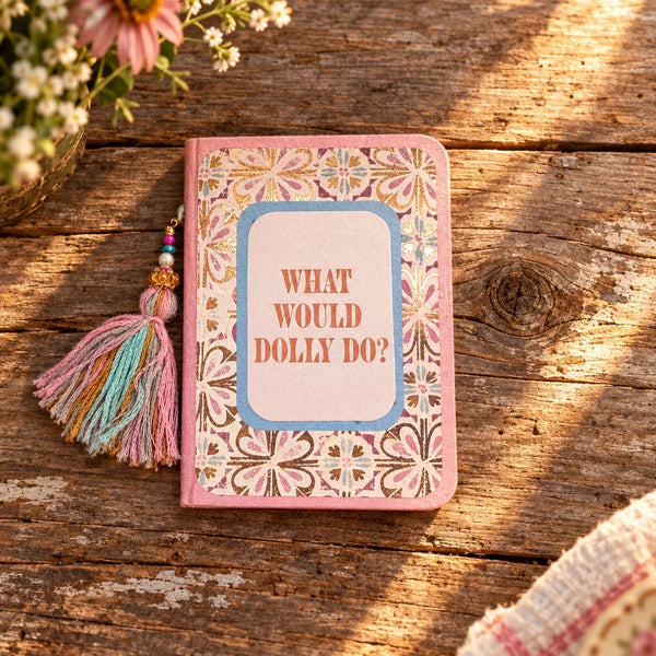 A small handmade journal or notebook with a pink border, floral patterned cover, and a tassel on the left side. The center features a light blue rounded rectangle with the text 'WHAT WOULD DOLLY DO?' in pink.