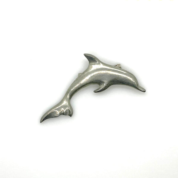 A vintage pewter dolphin brooch shown flat against a plain, off-white background. The brooch features a minimalist, sleek silver-tone design capturing a dolphin in a leaping arc, with a smooth matte finish, structured dorsal fin, flippers, and a detailed tail flipper.