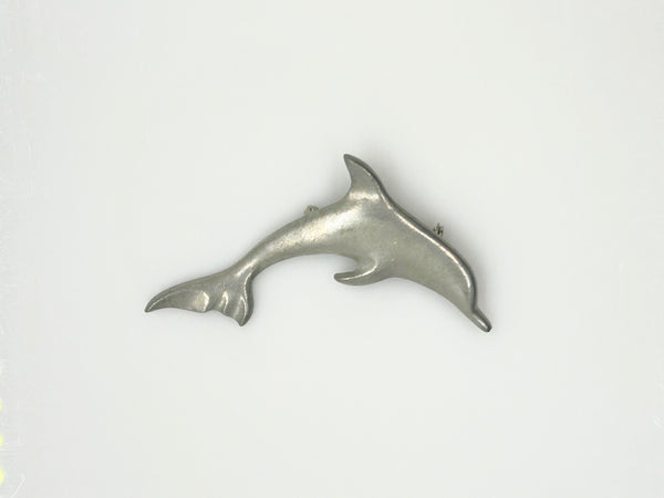 A vintage pewter dolphin brooch shown flat against a plain, off-white background. The brooch features a minimalist, sleek silver-tone design capturing a dolphin in a leaping arc, with a smooth matte finish, structured dorsal fin, flippers, and a detailed tail flipper.