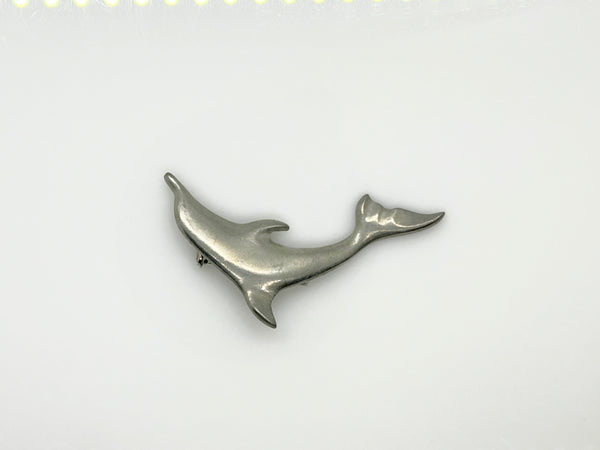 A vintage pewter dolphin brooch shown flat against a plain, off-white background. The brooch features a minimalist, sleek silver-tone design capturing a dolphin in a leaping arc, with a smooth matte finish, structured dorsal fin, flippers, and a detailed tail flipper.
