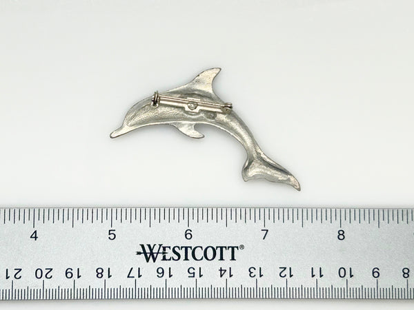 A vintage pewter dolphin brooch shown flat against a plain, off-white background. The brooch features a minimalist, sleek silver-tone design capturing a dolphin in a leaping arc, with a smooth matte finish, structured dorsal fin, flippers, and a detailed tail flipper.