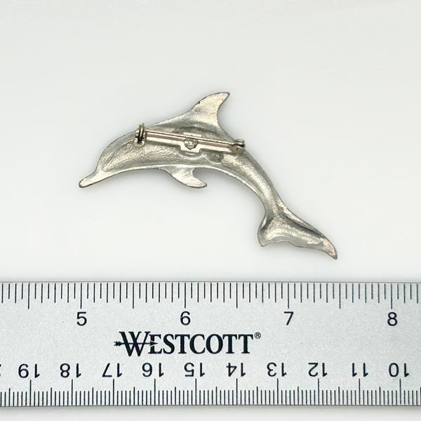 A vintage pewter dolphin brooch shown flat against a plain, off-white background. The brooch features a minimalist, sleek silver-tone design capturing a dolphin in a leaping arc, with a smooth matte finish, structured dorsal fin, flippers, and a detailed tail flipper.
