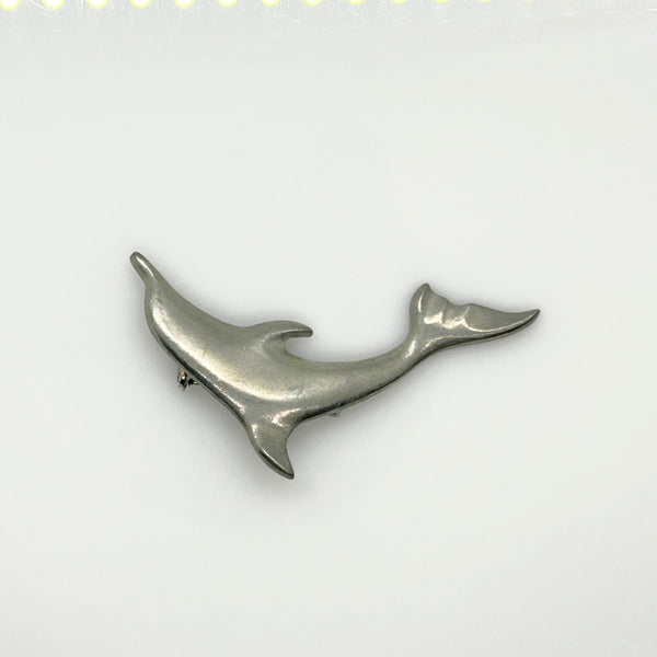 A vintage pewter dolphin brooch shown flat against a plain, off-white background. The brooch features a minimalist, sleek silver-tone design capturing a dolphin in a leaping arc, with a smooth matte finish, structured dorsal fin, flippers, and a detailed tail flipper.