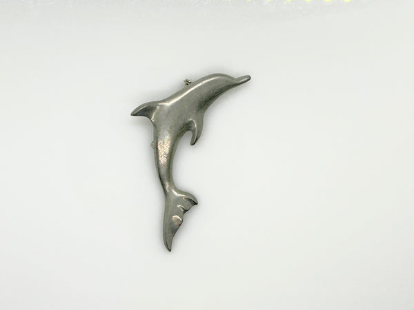 A vintage pewter dolphin brooch shown flat against a plain, off-white background. The brooch features a minimalist, sleek silver-tone design capturing a dolphin in a leaping arc, with a smooth matte finish, structured dorsal fin, flippers, and a detailed tail flipper.