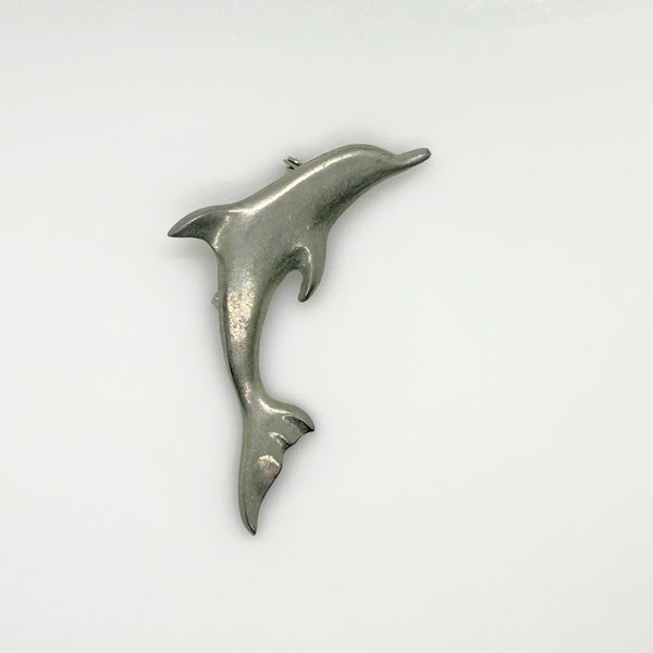A vintage pewter dolphin brooch shown flat against a plain, off-white background. The brooch features a minimalist, sleek silver-tone design capturing a dolphin in a leaping arc, with a smooth matte finish, structured dorsal fin, flippers, and a detailed tail flipper.