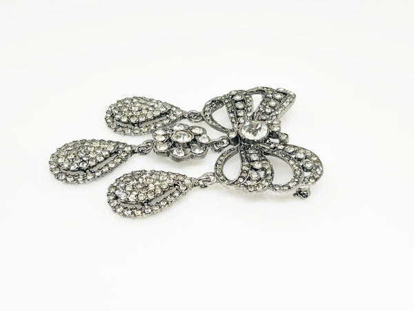 This exquisite piece of jewelry, crafted by Kenneth Jay Lane, is a shimmering testament to the exquisite beauty of the 18th century. It is dazzling, with crystal studded bows and gracefully dangling elaborate teardrops.The brooch is set in silver tone metal, its regal size measures 3.25" at its longest drop and 2" at its widest. The designer's signature is embossed on a raised oval at its back. Free Shipping