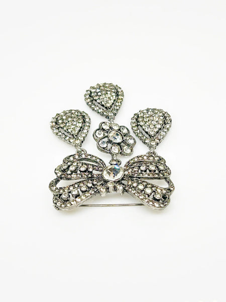 This exquisite piece of jewelry, crafted by Kenneth Jay Lane, is a shimmering testament to the exquisite beauty of the 18th century. It is dazzling, with crystal studded bows and gracefully dangling elaborate teardrops.The brooch is set in silver tone metal, its regal size measures 3.25" at its longest drop and 2" at its widest. The designer's signature is embossed on a raised oval at its back. Free Shipping