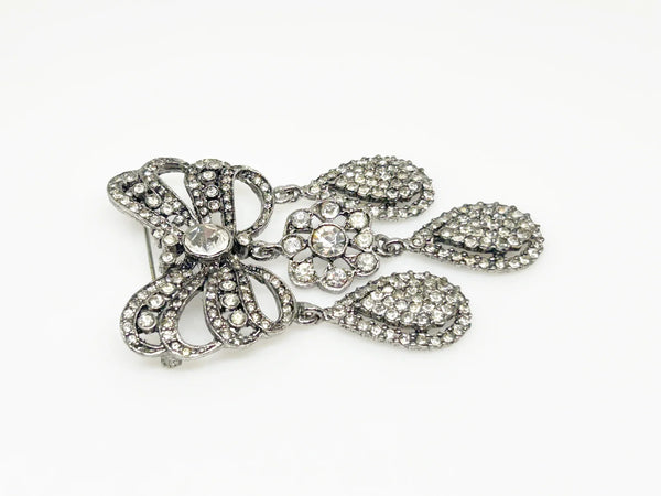 This exquisite piece of jewelry, crafted by Kenneth Jay Lane, is a shimmering testament to the exquisite beauty of the 18th century. It is dazzling, with crystal studded bows and gracefully dangling elaborate teardrops.The brooch is set in silver tone metal, its regal size measures 3.25" at its longest drop and 2" at its widest. The designer's signature is embossed on a raised oval at its back. Free Shipping